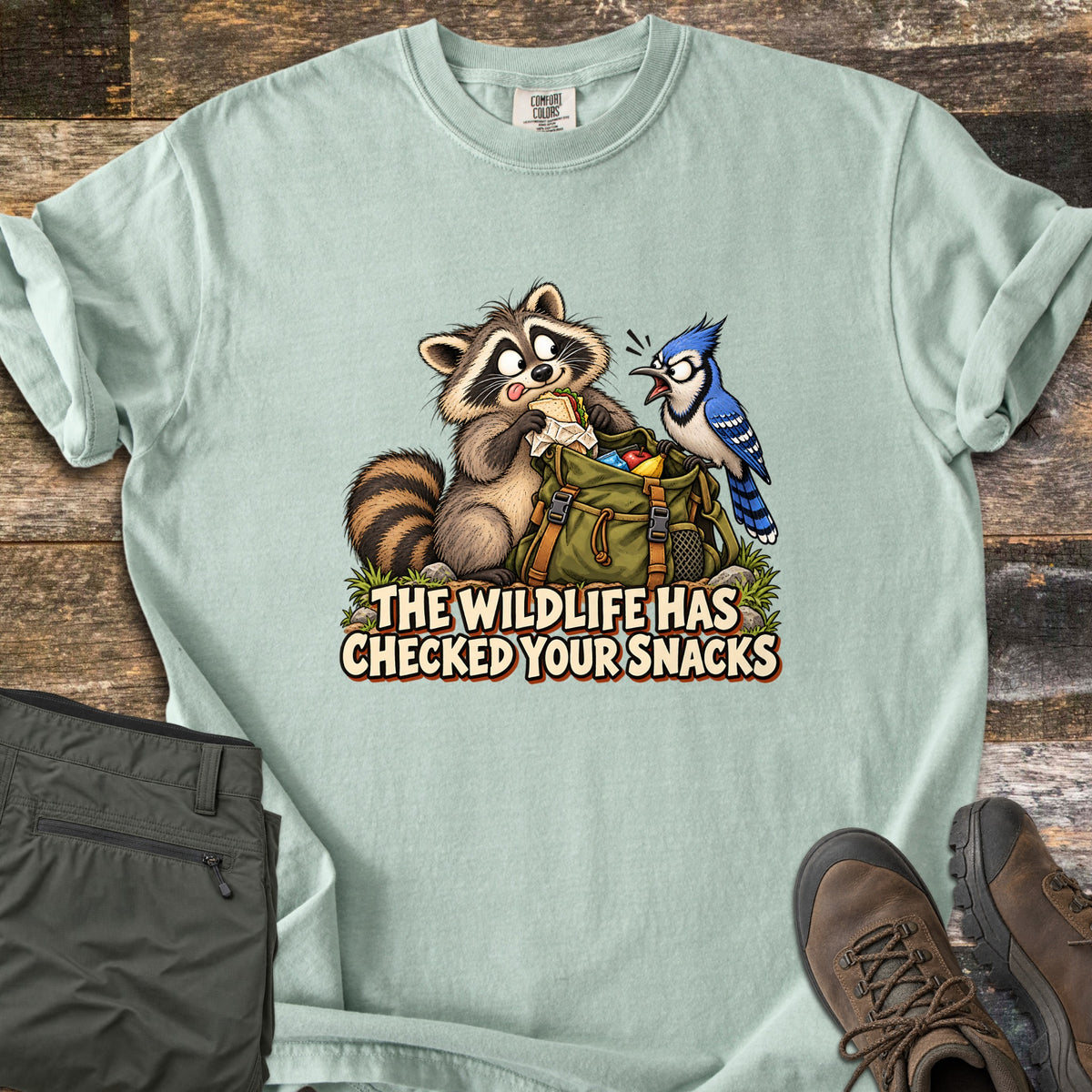 The Wildlife Has Checked Your Snacks Comfort Colors T-shirt in Bay, laid flat. A cartoon raccoon eats a sandwich from a green backpack while a startled blue jay squawks at it, above the words The Wildlife Has Checked Your Snacks. Olive hiking pants and brown boots lie on weathered wood.