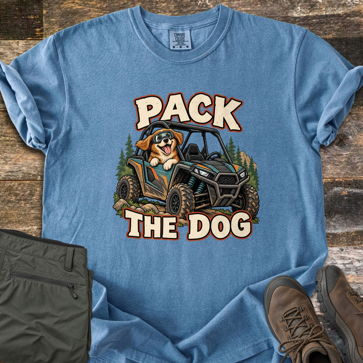 Pack the Dog Comfort Colors T-shirt in Blue Jean, laid flat. A cartoon dog in goggles rides in a muddy off-road side-by-side among pine trees, between the words Pack The Dog. Sleeves rolled, on weathered wood beside olive hiking pants and brown hiking boots.