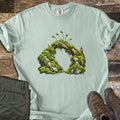 Moss Arch Comfort Colors T-shirt in Bay, laid flat. An illustrated arch of moss-covered rocks joined by a weathered branch, with slender moss sporophytes sprouting on top; no words. Sleeves are rolled, on a wood background with olive pants and brown hiking boots.