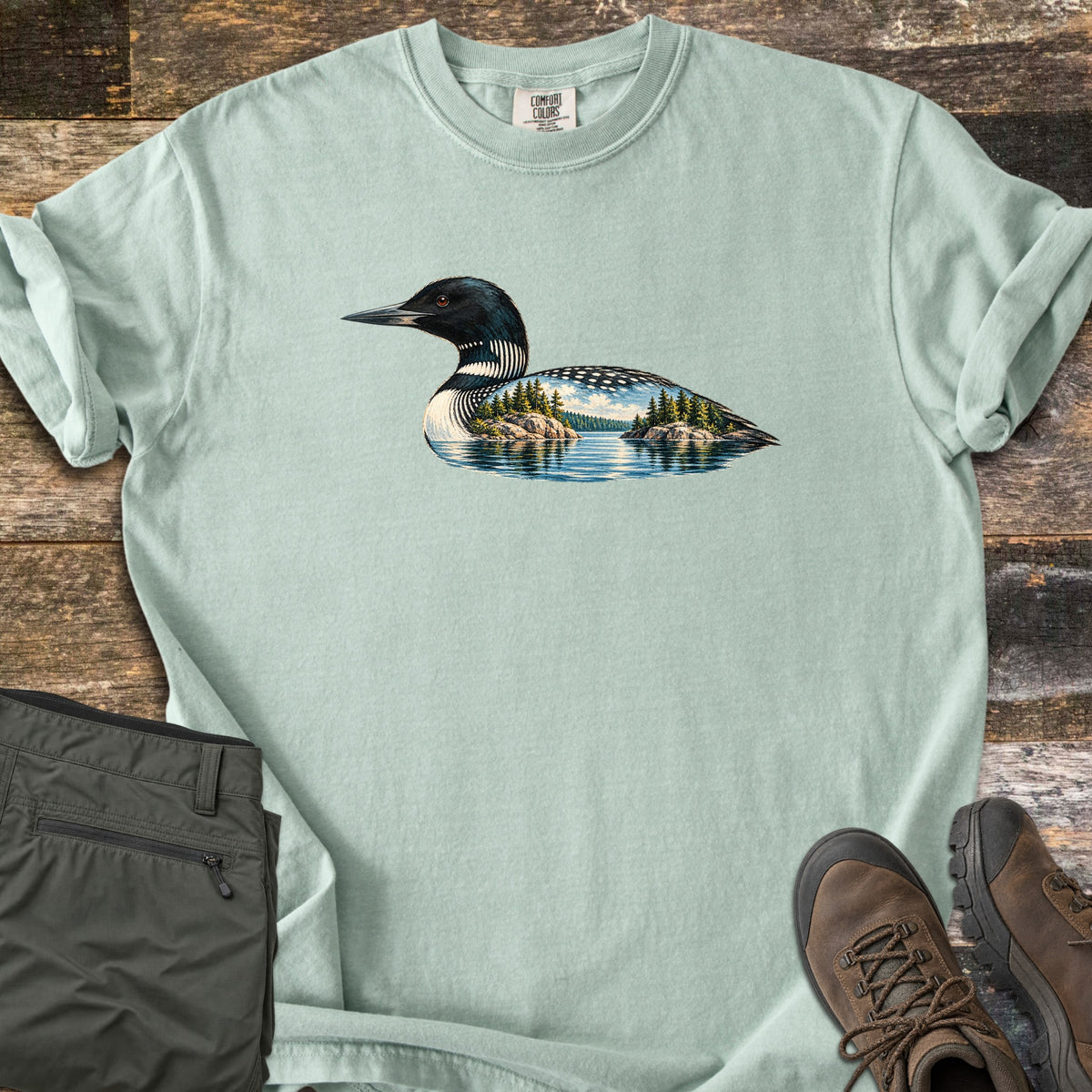 Loon Lake Comfort Colors T-shirt in Bay, laid flat. A common loon in black and white plumage whose body holds a lake scene with pine-topped rocky islands, blue sky and reflections. Olive hiking pants and brown hiking boots sit beside it on weathered wood planks.