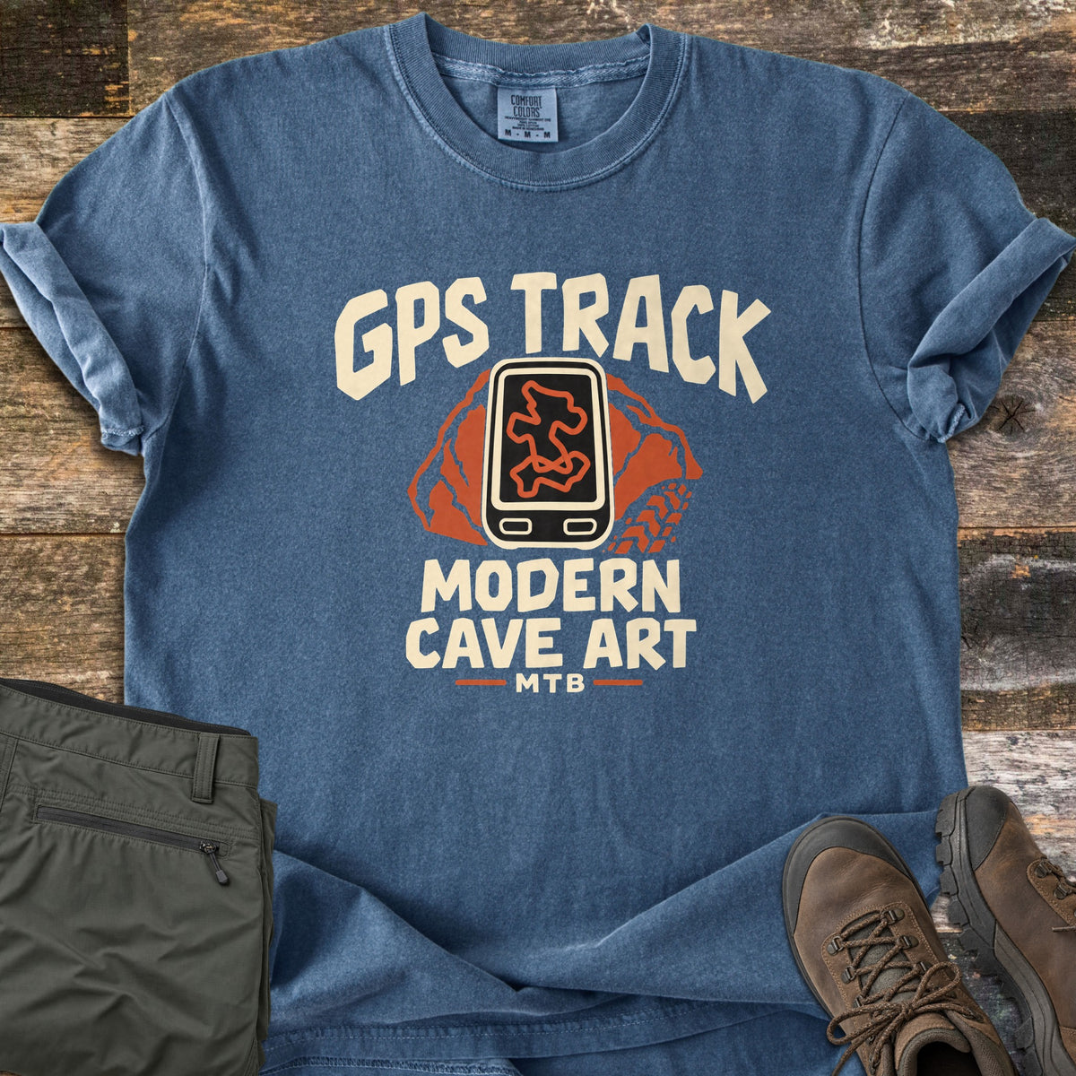 GPS Track Modern Cave Art Comfort Colors T-shirt in Denim, laid flat. Cream letters read GPS Track above a black GPS unit showing an orange trail route on an orange rock with a tire track, then Modern Cave Art and MTB. Folded olive pants and brown hiking boots lie on weathered wood.