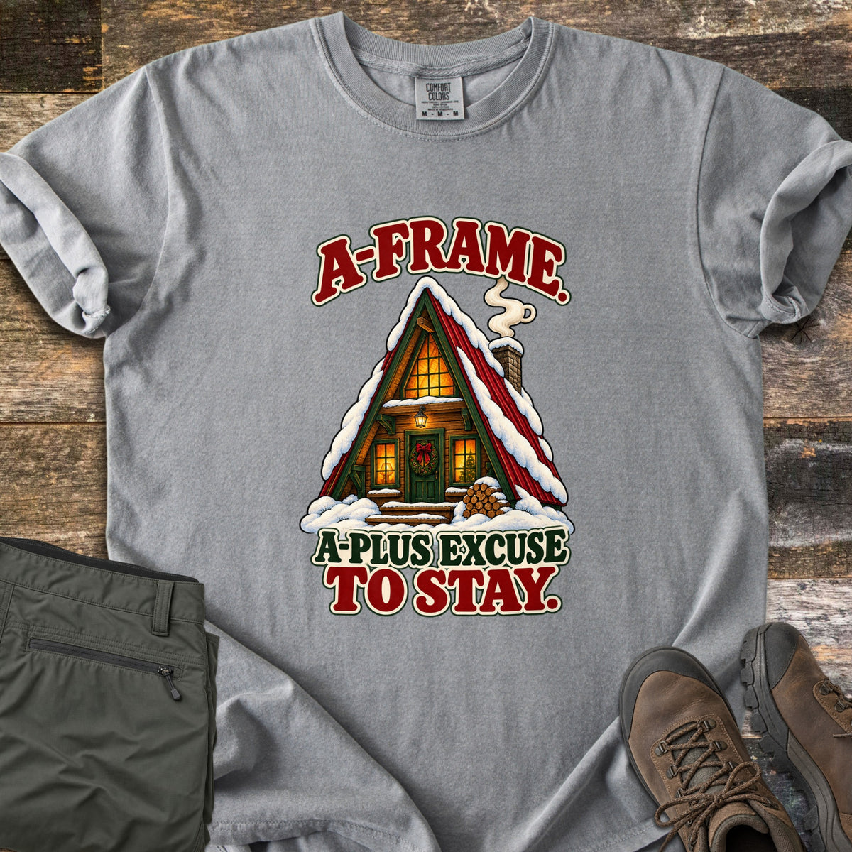 A-Frame A-Plus Excuse to Stay Comfort Colors T-shirt in Grey, laid flat. A snowy red-roofed A-frame cabin with glowing windows, a wreathed green door, smoking chimney and stacked firewood, between the words A-Frame. A-Plus Excuse to Stay. Olive pants and brown boots lie on weathered wood.