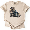Raccoon Night Creek Comfort Colors T-shirt in Sand, laid flat. A realistic raccoon sits looking to the side, its body filled with a moonlit creek winding through pine forest and mountains, and pine trees in its ringed tail. No words. Cuffed sleeves on a white background.