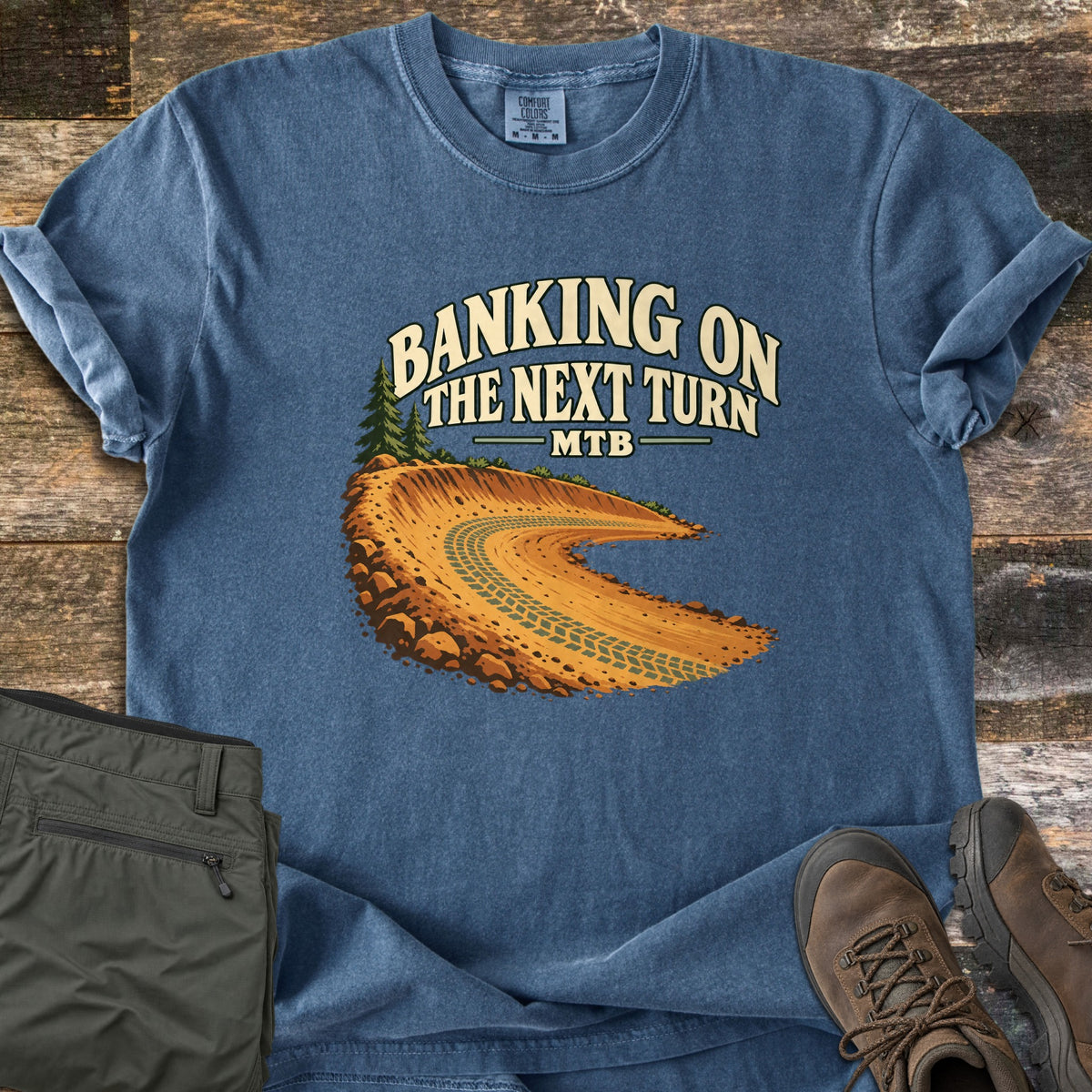 Banking on the Next Turn Comfort Colors T-shirt in Denim, laid flat. A dirt berm with tire tracks curves beneath pine trees, under the words Banking on the Next Turn MTB. Sleeves are rolled; olive hiking pants and brown hiking boots sit beside it on weathered wood.
