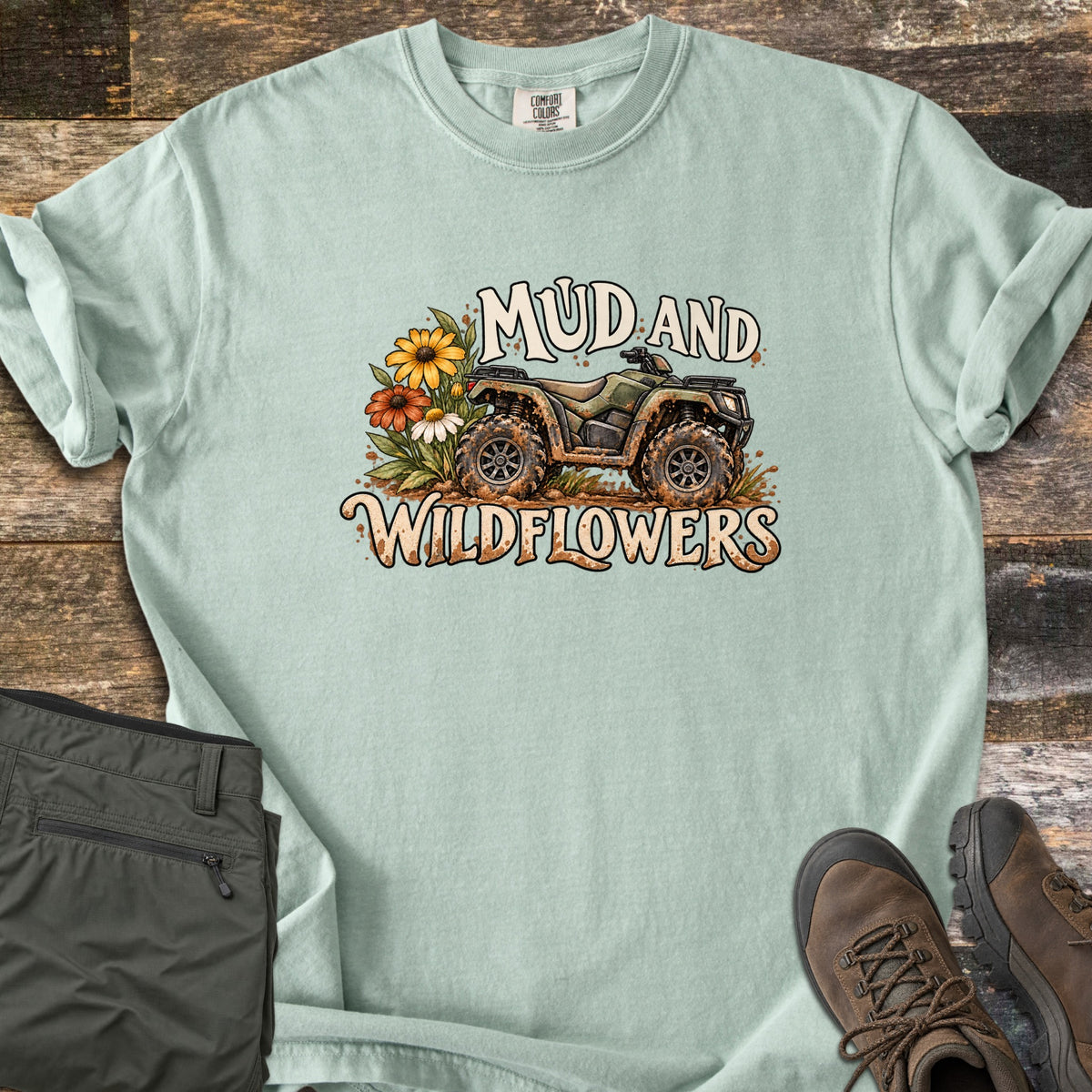 Mud and Wildflowers Comfort Colors T-shirt in Bay, laid flat. A mud-splattered green ATV beside yellow, orange and white wildflowers, with the words Mud and Wildflowers above and below. Folded olive pants and brown hiking boots sit on a weathered wood background.