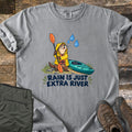 Extra River Comfort Colors T-shirt in Grey, laid flat. A cartoon otter in a yellow raincoat and life vest holds an orange paddle beside a teal kayak as raindrops fall, above the words Rain Is Just Extra River. Olive pants and brown hiking boots lie on a wood floor.