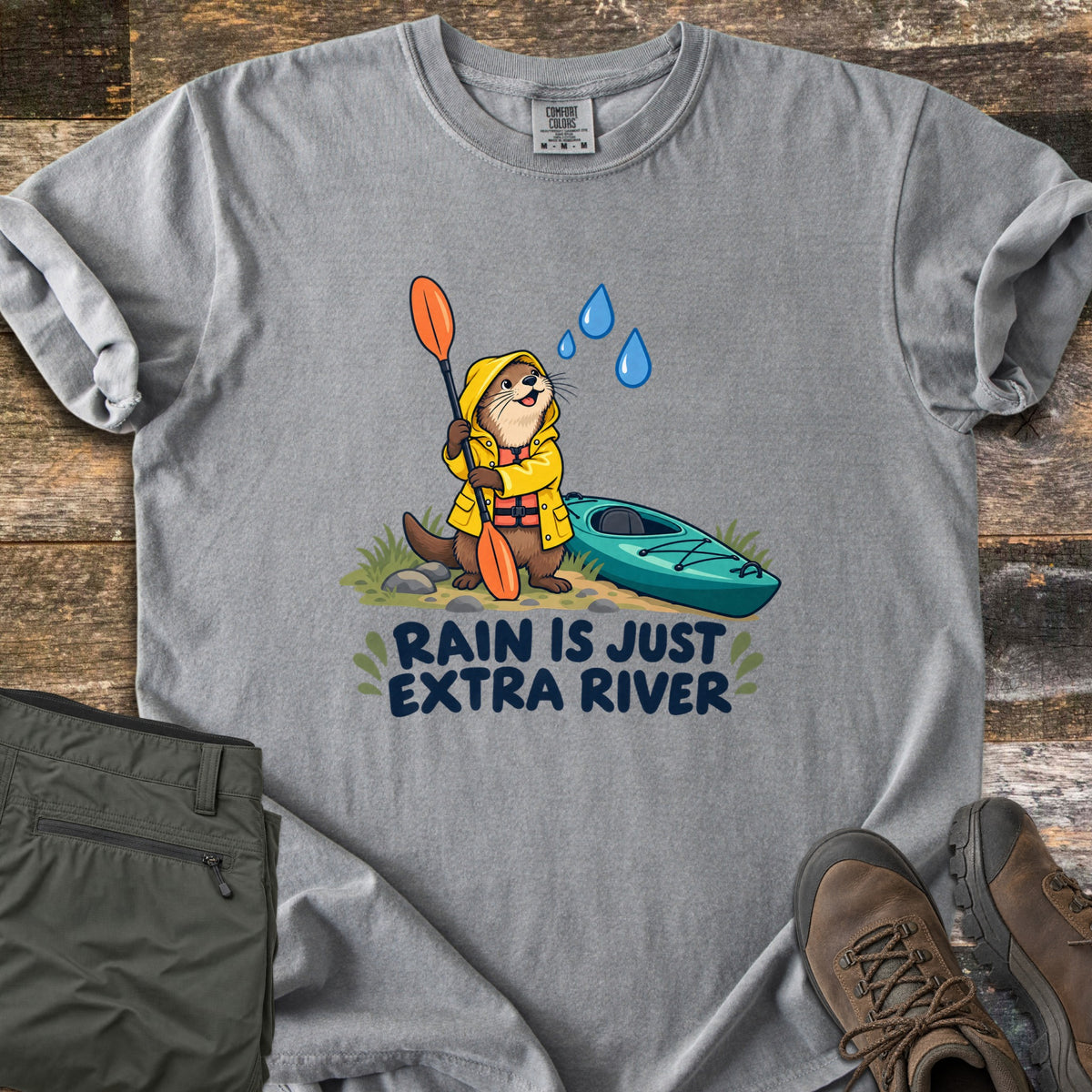 Extra River Comfort Colors T-shirt in Grey, laid flat. A cartoon otter in a yellow raincoat and life vest holds an orange paddle beside a teal kayak as raindrops fall, above the words Rain Is Just Extra River. Olive pants and brown hiking boots lie on a wood floor.