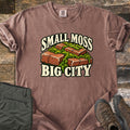 Small Moss Big City Comfort Colors T-shirt in Espresso, laid flat. Red bricks with bright green moss growing between and over them, with the words Small Moss Big City in cream lettering. Sleeves are rolled, on a wood floor beside folded olive pants and brown hiking boots.