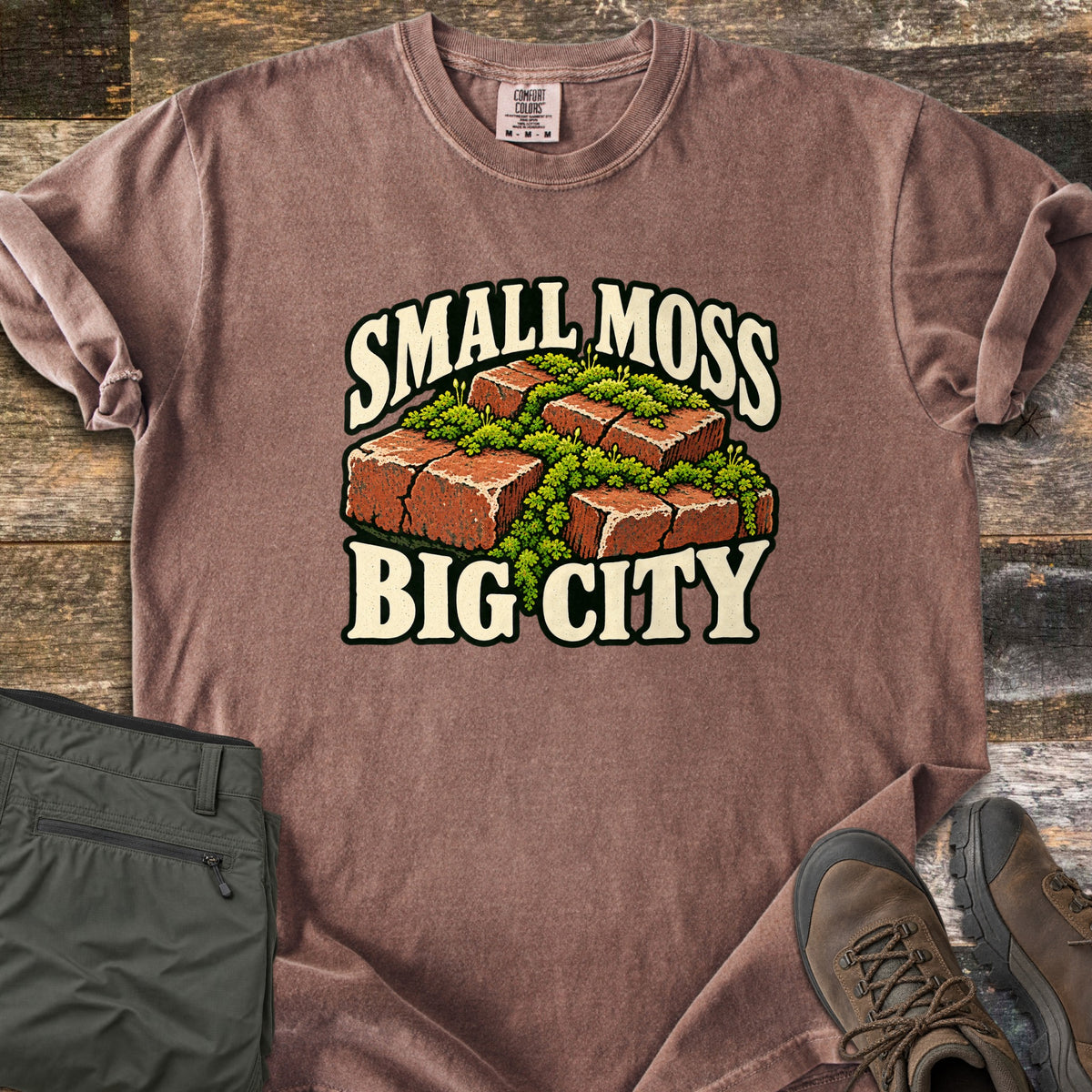 Small Moss Big City Comfort Colors T-shirt in Espresso, laid flat. Red bricks with bright green moss growing between and over them, with the words Small Moss Big City in cream lettering. Sleeves are rolled, on a wood floor beside folded olive pants and brown hiking boots.