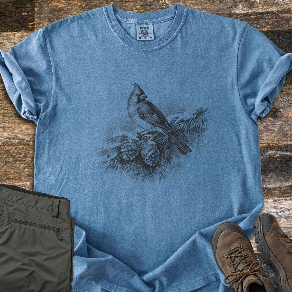 Cardinal On A Snowy Bough Comfort Colors T-shirt in Blue Jean, laid flat. A grayscale drawing of a cardinal perched on a snow-covered pine branch with two pinecones, with no words. Sleeves rolled, on weathered wood planks beside olive hiking pants and brown hiking boots.