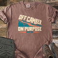 Off Camber On Purpose Comfort Colors T-shirt in Espresso, laid flat. Cream block letters read Off Camber On Purpose around a teal mountain slope with an orange zigzag trail, with MTB in teal below. Olive hiking pants and brown boots sit beside it on weathered wood planks.