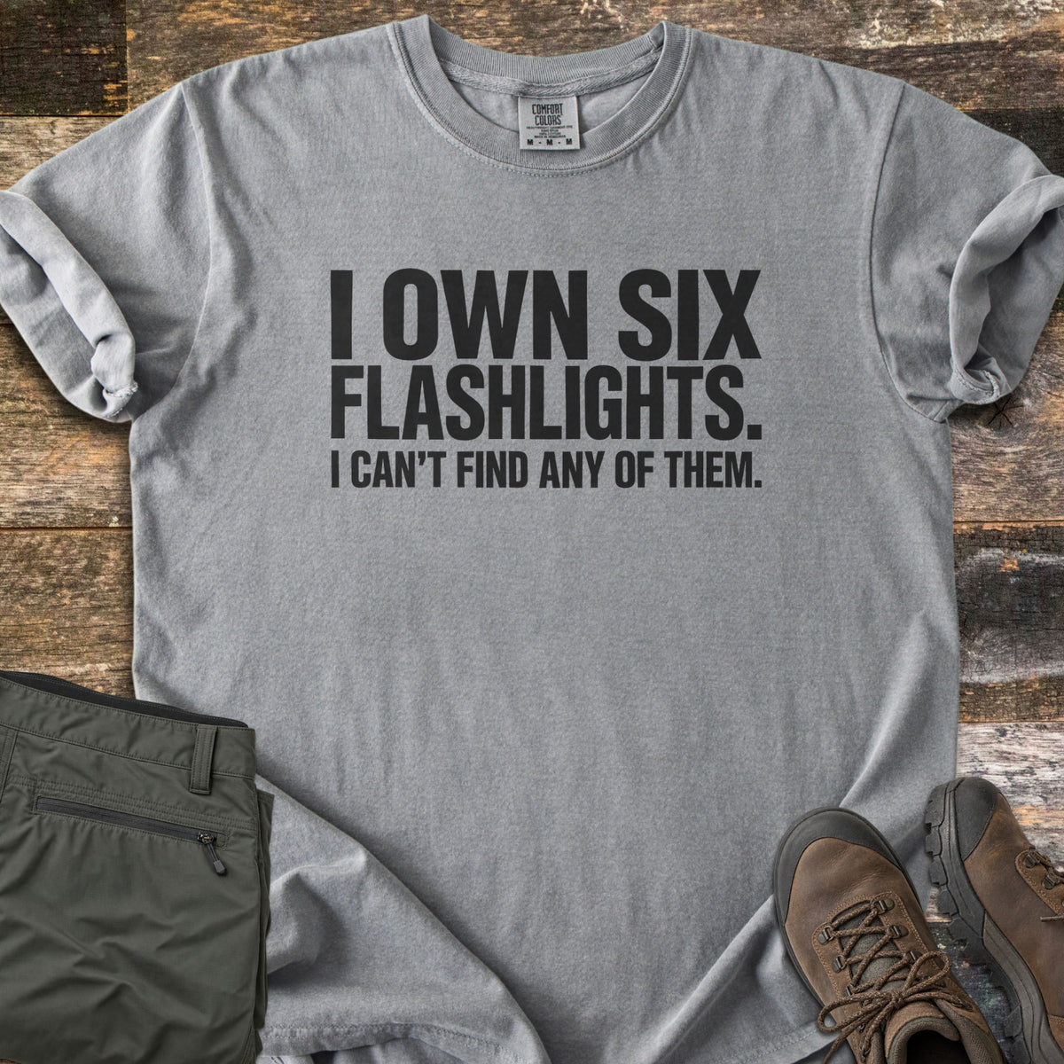 Six Flashlights Comfort Colors T-shirt in Grey, laid flat. Bold black text reads I OWN SIX FLASHLIGHTS. I CAN'T FIND ANY OF THEM. The shirt has rolled sleeves and lies on weathered wood beside folded olive cargo pants and brown hiking boots.