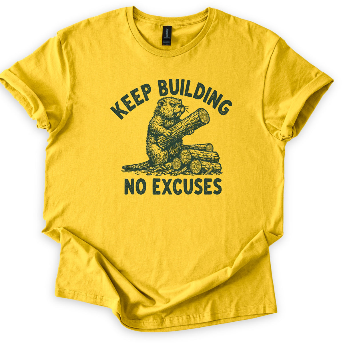 Keep Building Beaver Comfort Colors T-shirt in Daisy, laid flat. Dark green sketch-style artwork of a beaver hugging a log while sitting on a pile of cut logs, with Keep Building arched above and No Excuses below. Sleeves are rolled on a white background.