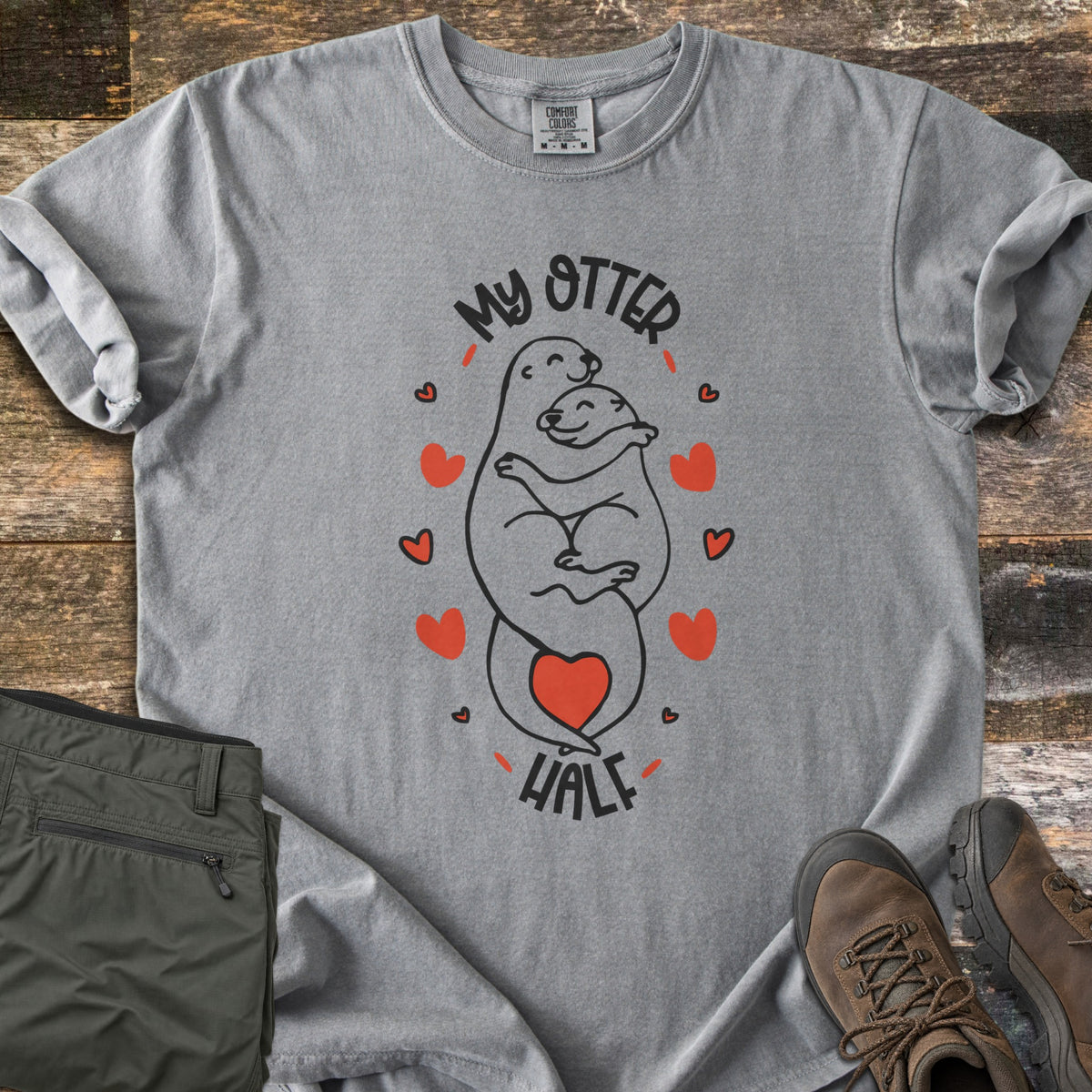 My Otter Half Comfort Colors T-shirt in Grey, laid flat. Two smiling cartoon otters hug in black line art, ringed by red hearts with a red heart at their tails, between the words My Otter Half. Folded olive pants and brown hiking boots sit beside it on weathered wood.