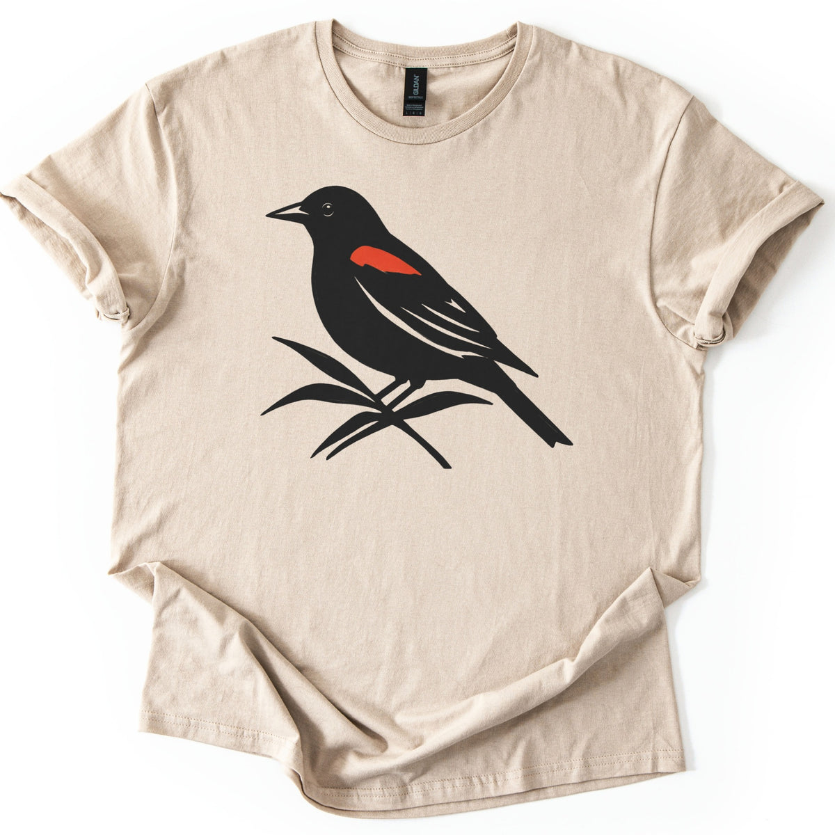 Red Winged Blackbird Comfort Colors T-shirt in Sand, laid flat. A black silhouette of a red-winged blackbird with a red shoulder patch, perched on slender leaves, with no words. The shirt has rolled sleeves and lies on a white background.
