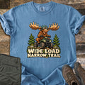 Wide Load Narrow Trail Comfort Colors T-shirt in Blue Jean, laid flat. A cartoon moose with huge antlers, a helmet and goggles rides a green ATV between pine trees above the words Wide Load Narrow Trail. Olive pants and brown hiking boots lie beside it on weathered wood.