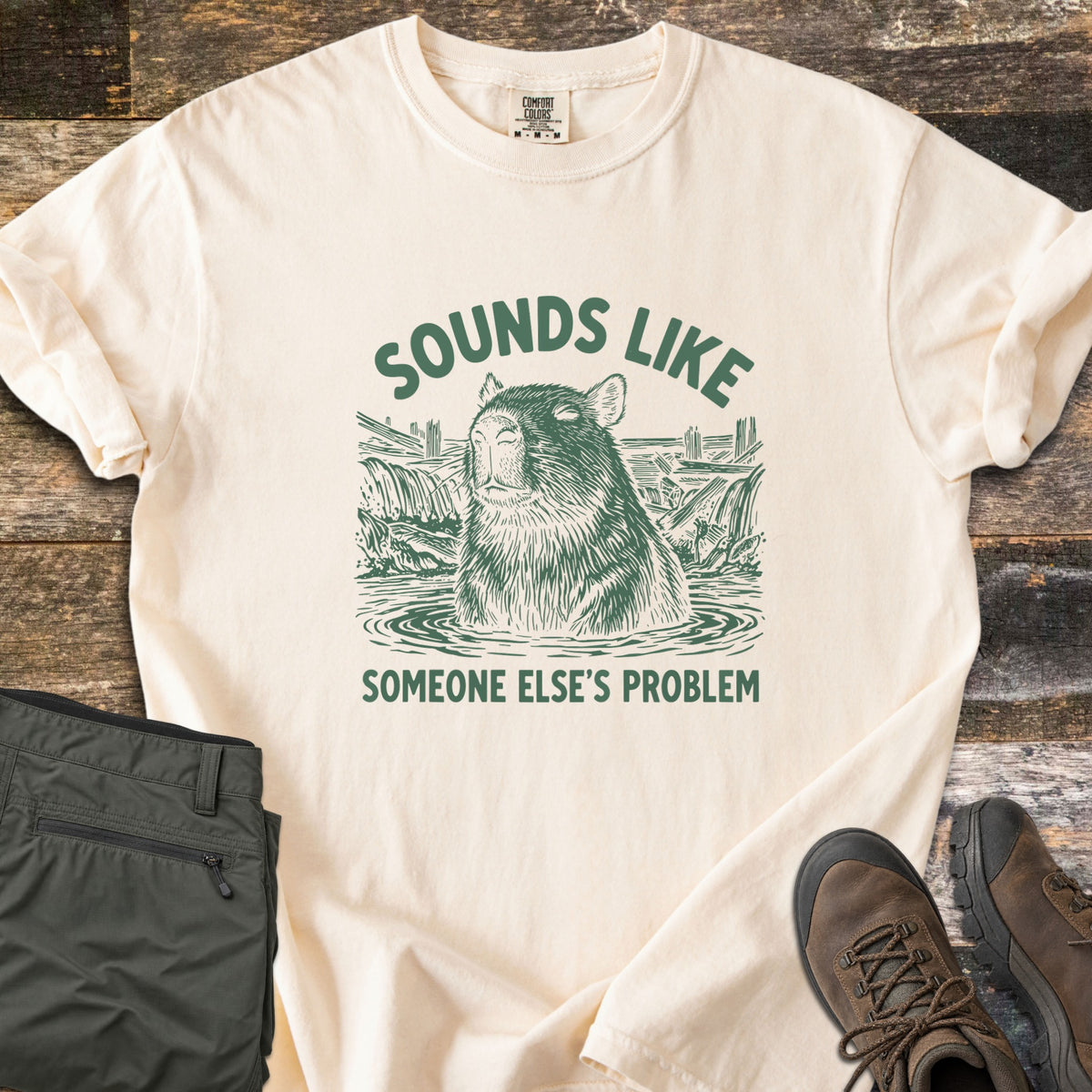 Sounds Like Someone Else's Problem Comfort Colors T-shirt in Ivory, laid flat. Green sketch-style art of a capybara sitting in a pond with eyes closed, under the words Sounds Like and above Someone Else's Problem. Olive pants and brown hiking boots lie on a dark wood surface.