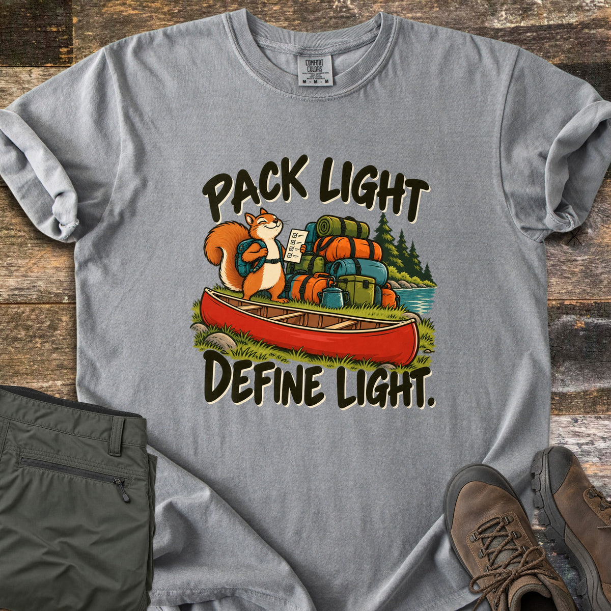Pack Light Define Light Comfort Colors T-shirt in Grey, laid flat. A smiling cartoon squirrel with a backpack holds a checklist beside a red canoe piled with gear bags, between the words Pack Light and Define Light. Olive pants and brown hiking boots sit on weathered wood.
