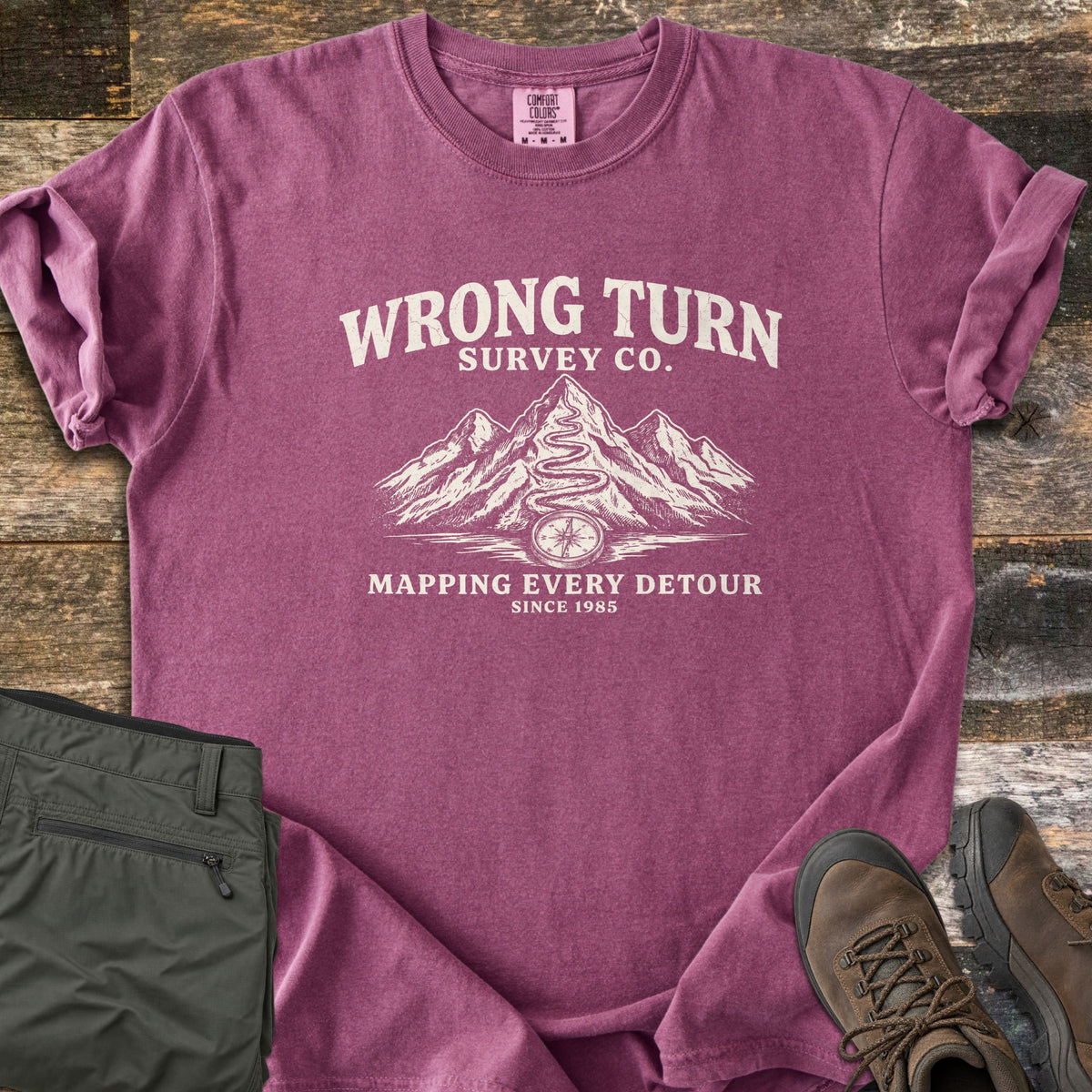 Wrong Turn Survey Co Comfort Colors T-shirt in Berry, laid flat. Cream print of the words Wrong Turn Survey Co. above snowy mountains with a winding trail leading to a compass, then Mapping Every Detour, Since 1985. Shown on wood boards with olive pants and brown hiking boots.