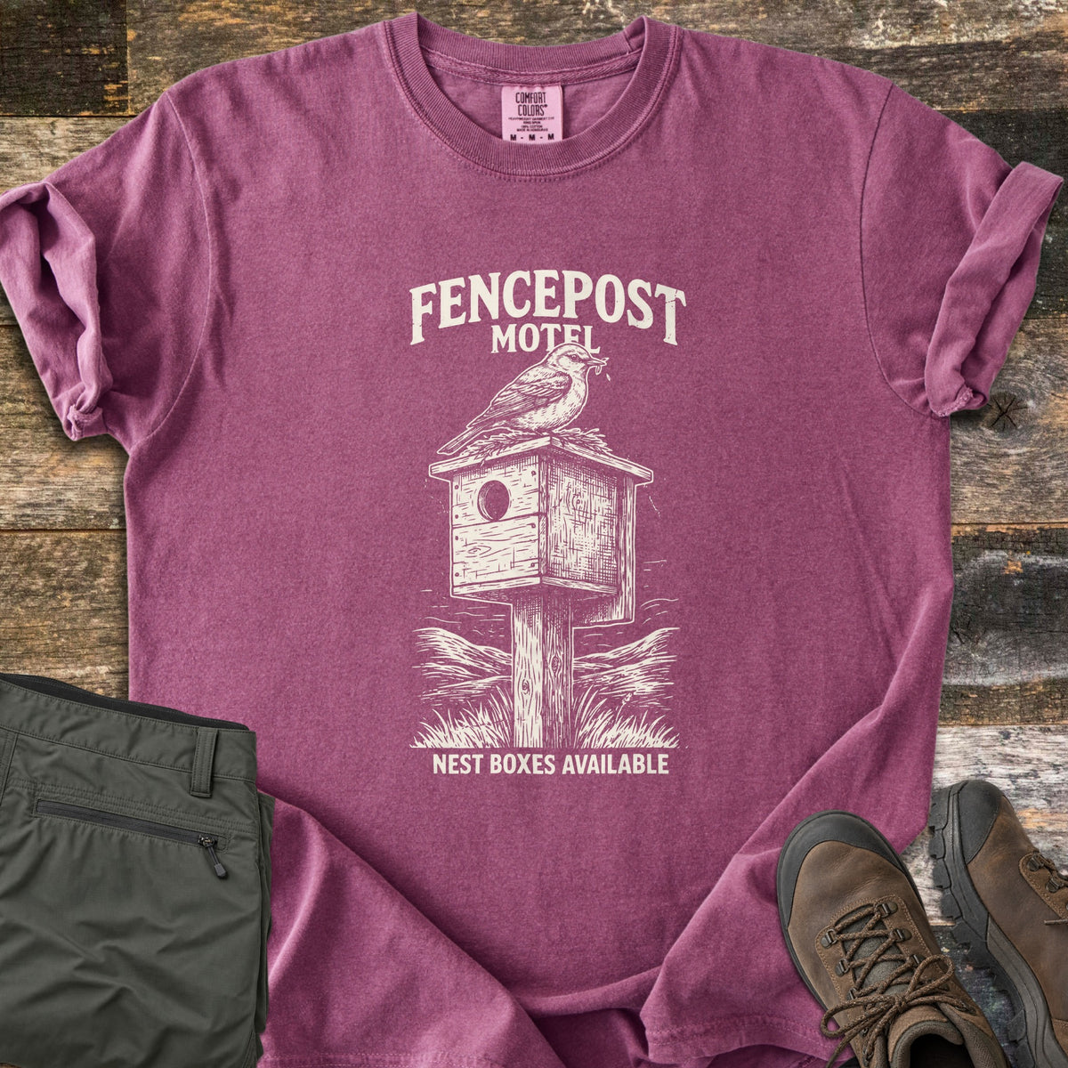 Fencepost Motel Comfort Colors T-shirt in Berry, laid flat. A cream sketch of a bird perched on a wooden nest box atop a post in tall grass, with the words Fencepost Motel above and Nest Boxes Available below. Sleeves are rolled on a wood plank surface beside olive pants and brown hiking boots.
