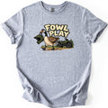 Fowl Play Comfort Colors T-shirt in Sport Grey, laid flat. A cartoon Canada goose carries a green motocross helmet in its beak past a green ATV and pine trees, below the words Fowl Play. The shirt lies on a white background with cuffed sleeves.