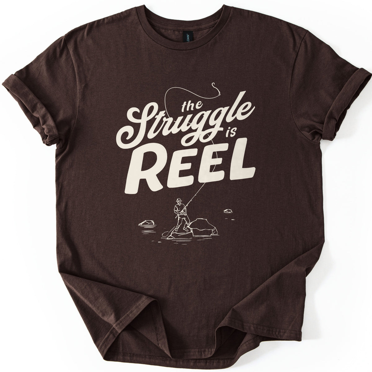 Struggle Is Reel Angler Comfort Colors T-shirt in Dark Chocolate, laid flat. Cream text reads the Struggle is REEL, with a fishing line arcing over the words to a hook. Below, a line drawing of an angler in a hat casts from a rock in the water. Sleeves cuffed, on a white background.