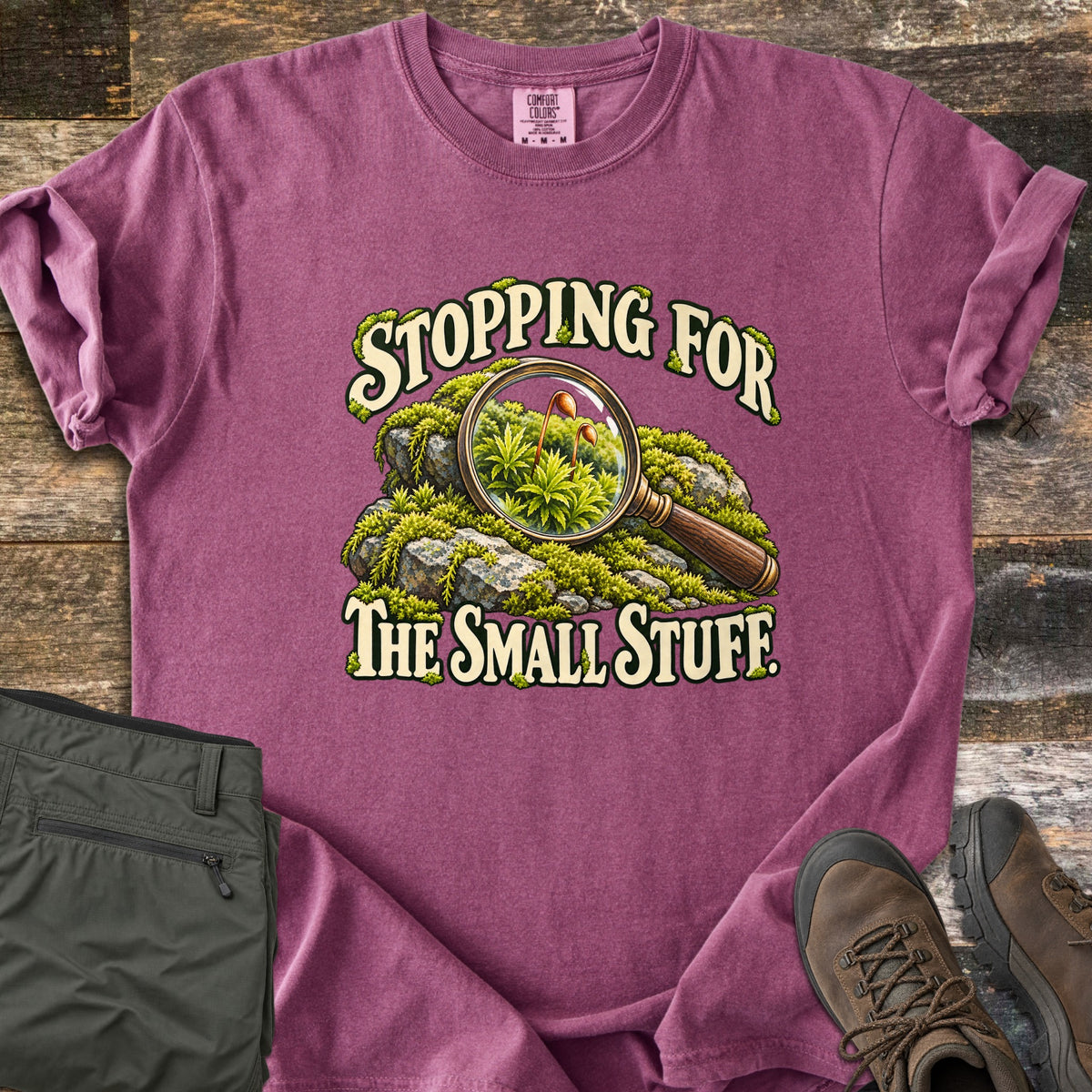Stopping for the Small Stuff Comfort Colors T-shirt in Berry, laid flat. A magnifying glass rests on mossy rocks, showing tiny moss sprouts, with the words Stopping for the Small Stuff. On rustic wood beside folded olive hiking pants and brown hiking boots.