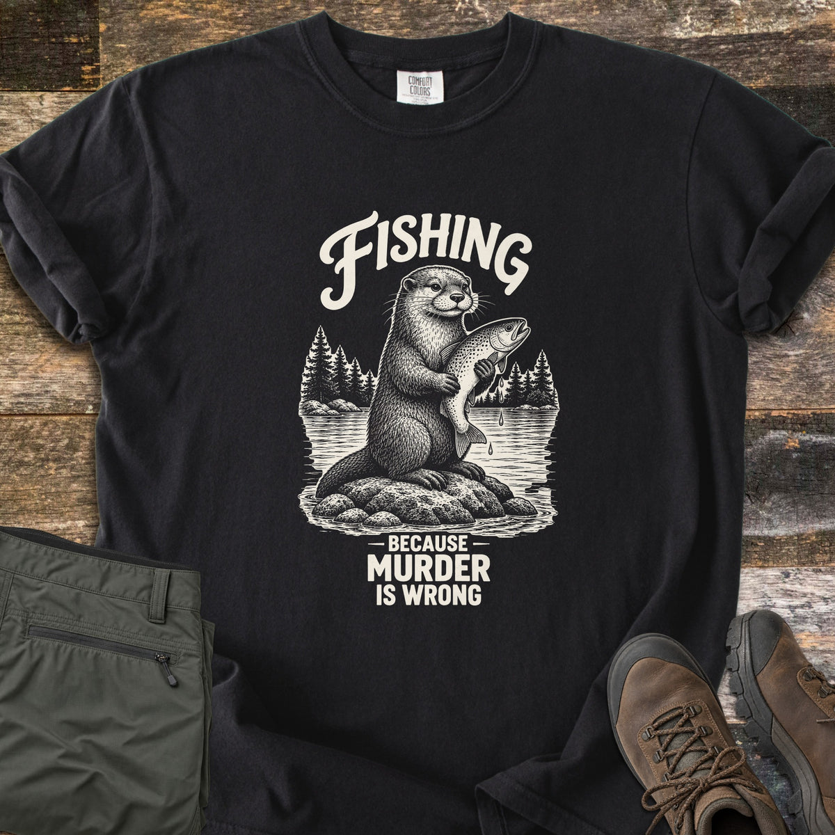 Fishing Murder Otter Comfort Colors T-shirt in Black, laid flat. Cream ink artwork of an otter on a rock holding a dripping trout before a pine-lined lake, with Fishing above and Because Murder Is Wrong below. Olive pants and brown hiking boots sit on weathered wood.