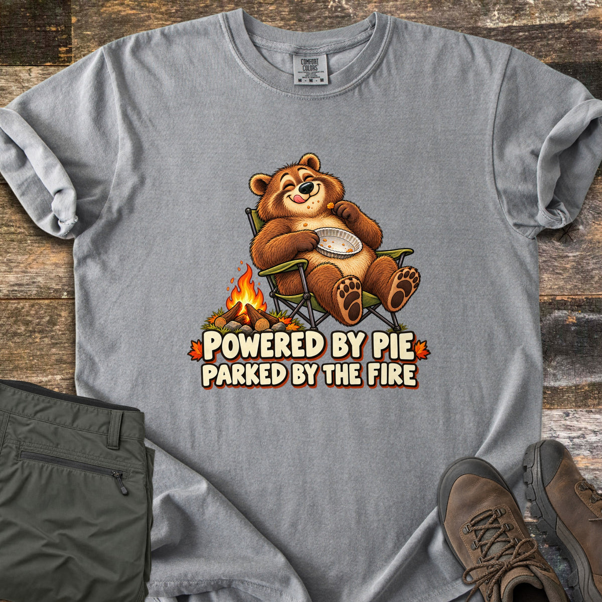 Powered by Pie Parked by the Fire Comfort Colors T-shirt in Grey, laid flat. A cartoon brown bear lounges in a green camp chair holding an empty pie tin beside a campfire, above the words Powered by Pie Parked by the Fire. Olive pants and brown hiking boots lie on weathered wood.