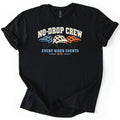No Drop Crew Comfort Colors T-shirt in Black, laid flat. Three mountain bike helmets in blue, cream and orange sit under the arched words No-Drop Crew, with Every Rider Counts and MTB below. The shirt has rolled sleeves on a white background.