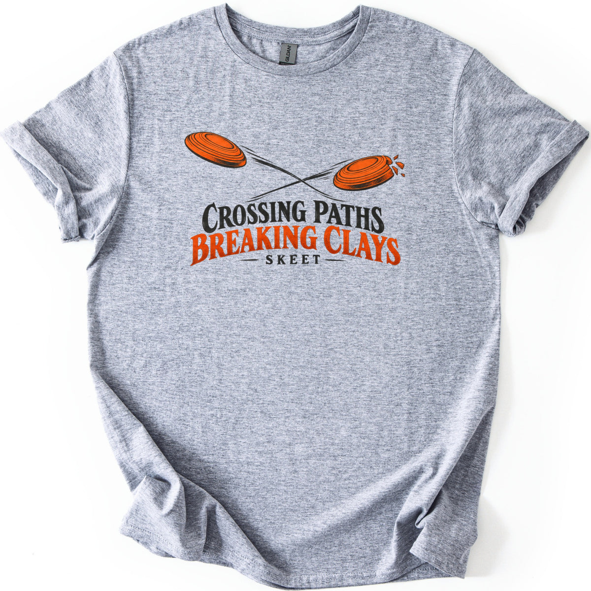 Crossing Paths Breaking Clays Comfort Colors T-shirt in Sport Grey, laid flat. Two orange clay targets fly on crossing paths, the right one breaking apart, above the words Crossing Paths Breaking Clays Skeet. The shirt lies on a white background with its sleeves cuffed.