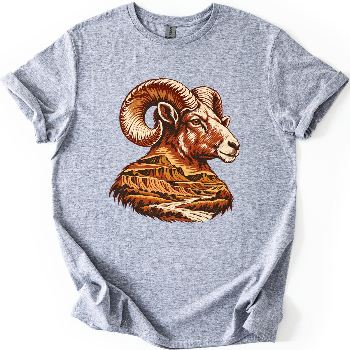 Bighorn Badlands Comfort Colors T-shirt in Sport Grey, laid flat. A bighorn ram's head in profile with curled horns, in rust, orange and cream, its neck filled with layered badlands buttes and a winding river. No words are printed. The shirt lies on white with cuffed sleeves.