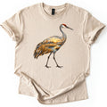 Crane Marsh Sunrise Comfort Colors T-shirt in Sand, laid flat. A walking sandhill crane with a red crown, its body filled with a painted marsh at sunrise: golden reeds, a winding stream and the sun over distant trees. No words. Cuffed sleeves on a white background.