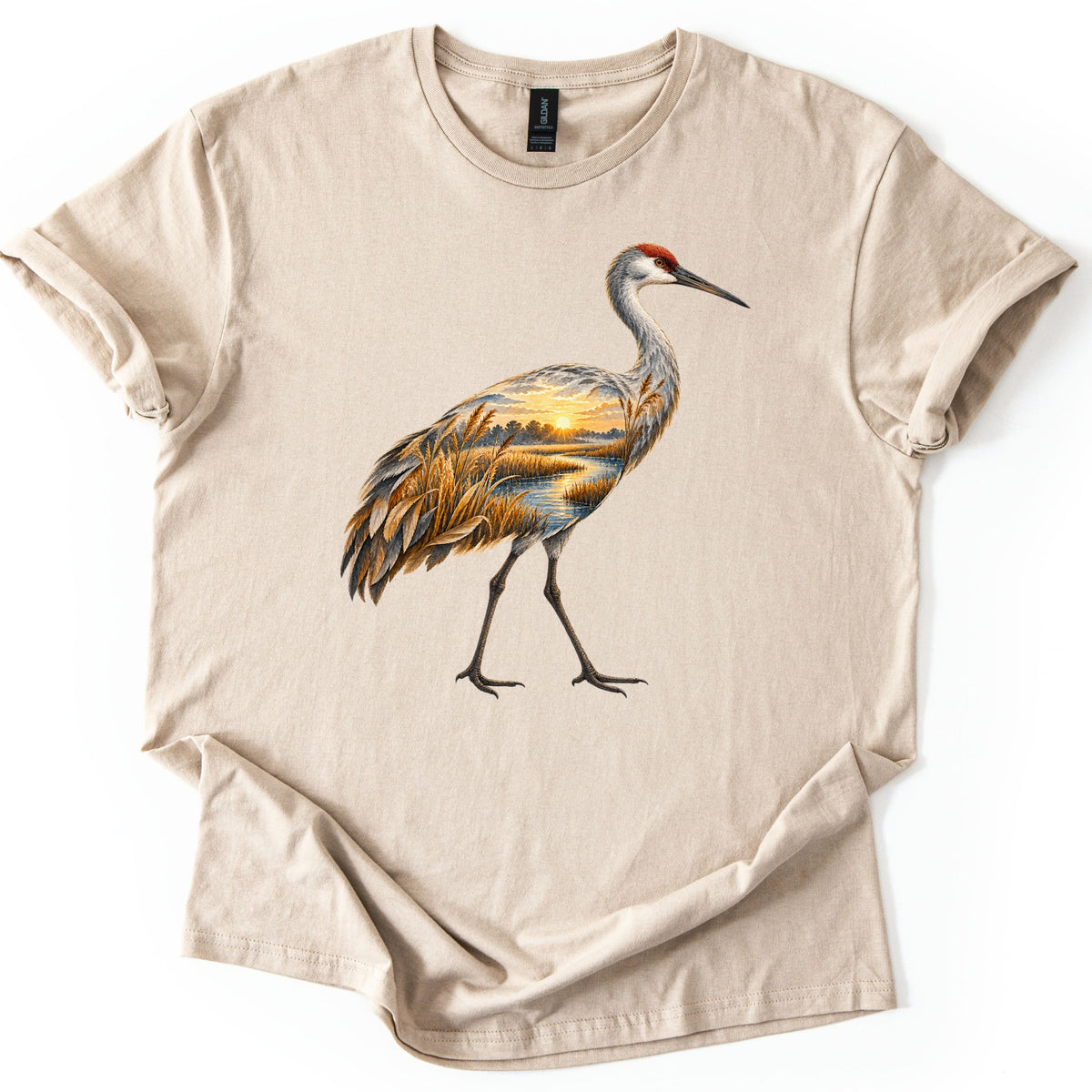 Crane Marsh Sunrise Comfort Colors T-shirt in Sand, laid flat. A walking sandhill crane with a red crown, its body filled with a painted marsh at sunrise: golden reeds, a winding stream and the sun over distant trees. No words. Cuffed sleeves on a white background.