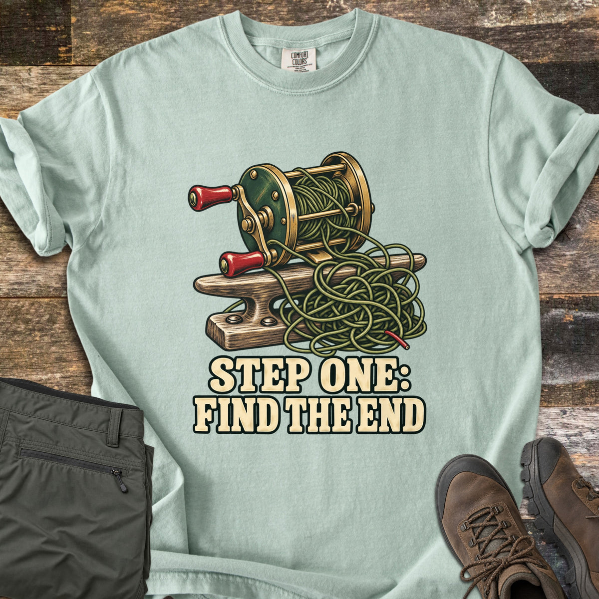 Step One: Find the End Comfort Colors T-shirt in Bay, laid flat. A vintage green and brass fishing reel with red handles sits on a wooden boat cleat, its line in a tangled pile, above the words Step One: Find the End. Shown on weathered wood with olive pants and brown hiking boots.
