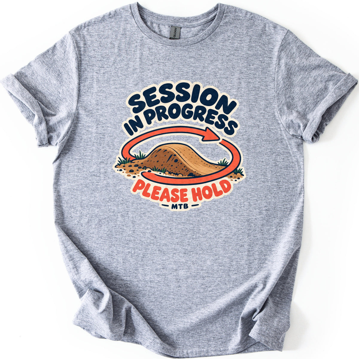 Session in Progress Comfort Colors T-shirt in Sport Grey, laid flat. A dirt jump mound with tufts of grass is circled by an orange looping arrow, with the words Session in Progress above and Please Hold MTB below. The sleeves are cuffed on a white background.