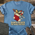 Santa Packed The Hooks Together Comfort Colors T-shirt in Blue Jean, laid flat. A red knit Christmas stocking holds fishing lures, a bobber and a rod handle tied with a cream ribbon above the words Santa Packed The Hooks Together. Olive pants and brown boots sit on a wood floor.