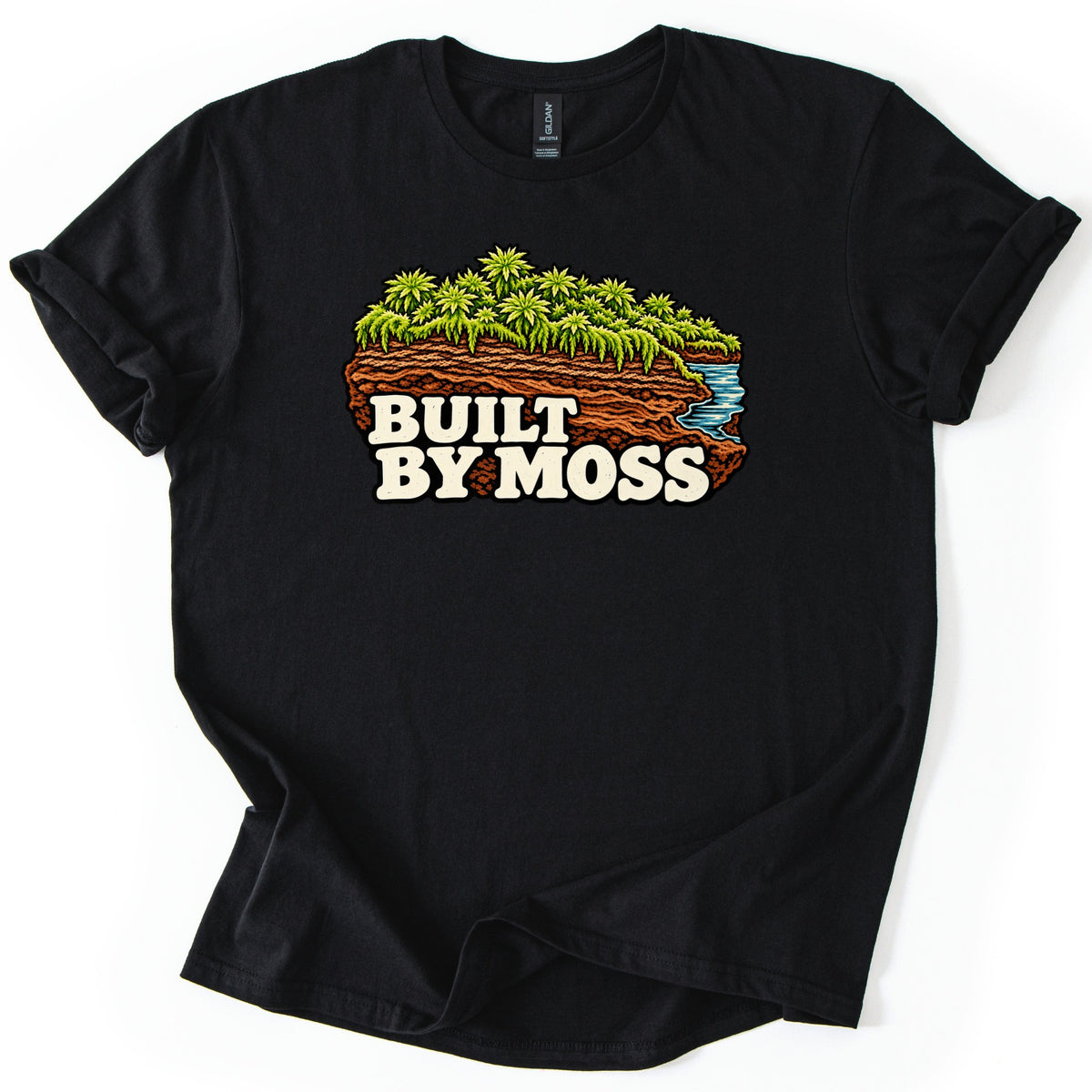 Built by Moss Comfort Colors T-shirt in Black, laid flat. A chunk of brown soil topped with bright green moss, with a small blue stream flowing off one edge, above the words Built By Moss in cream letters. The shirt has rolled sleeves and lies on a white background.