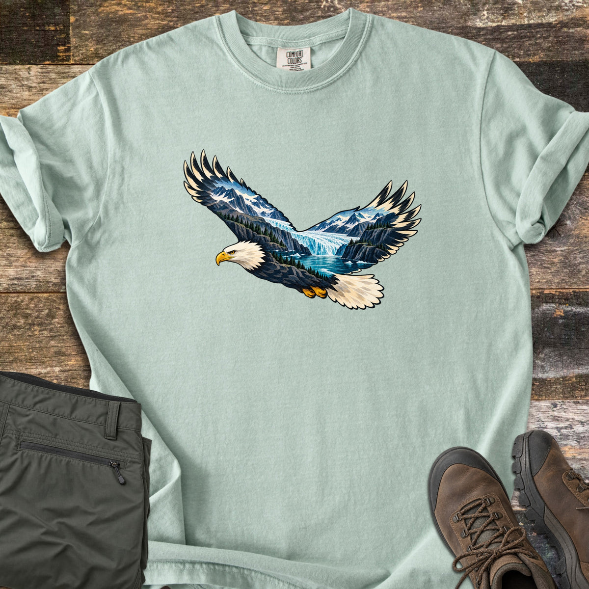 Eagle Glacier Valley Comfort Colors T-shirt in Bay, laid flat. A bald eagle in flight with a mountain glacier valley, waterfall, pine trees and blue lake shown inside its wings and body; no words. Olive cargo shorts and brown hiking boots sit beside it on weathered wood.