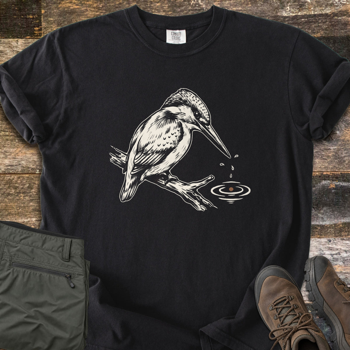Kingfisher Comfort Colors T-shirt in Black, laid flat. A cream ink sketch of a kingfisher perched on a branch, looking down at water drops falling from its beak into rippling water, with no words. Olive pants and brown hiking boots sit beside it on weathered wood.
