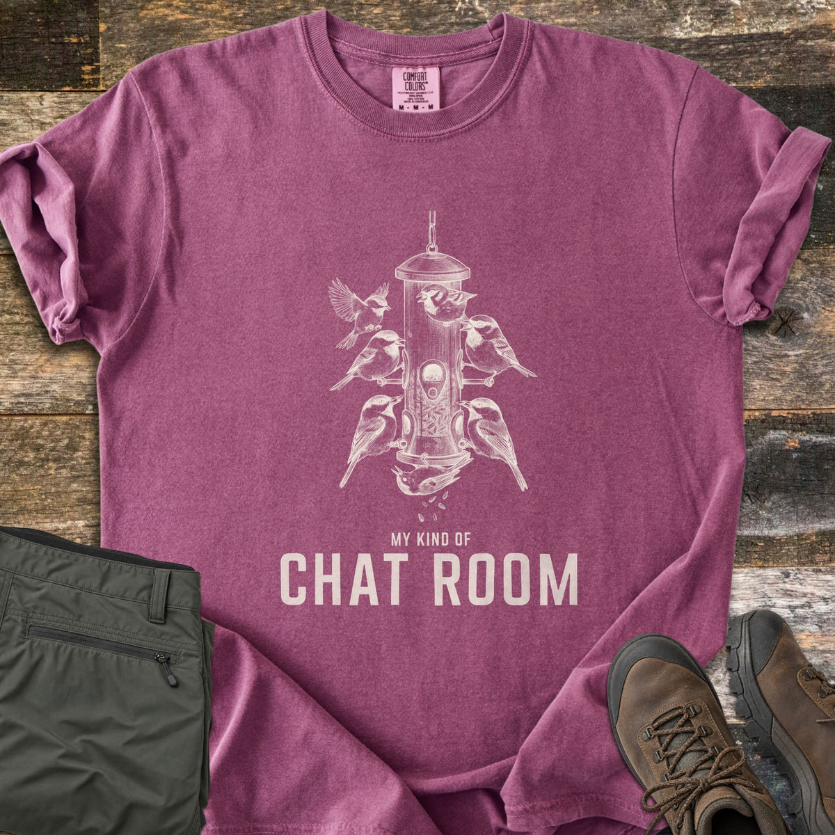 My Kind Of Chat Room Comfort Colors T-shirt in Berry, laid flat. A white sketch of chickadees perched around a hanging tube bird feeder, one upside down below it, above the words My Kind Of Chat Room. Olive hiking pants and brown boots lie beside it on weathered wood.