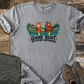 Same Boat Different Opinions Comfort Colors T-shirt in Grey, laid flat. A cartoon fox in a life vest points ahead while a calm bear paddles a green canoe on a lake with pine trees, above the words Same Boat Different Opinions. Olive pants and brown hiking boots lie on wood planks.