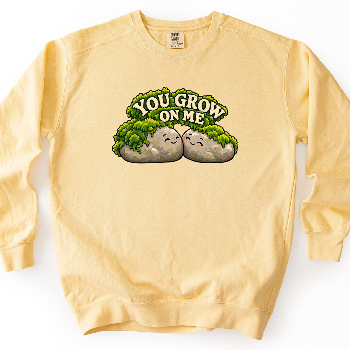You Grow on Me Comfort Colors crewneck sweatshirt in Butter, laid flat. Two smiling gray rocks covered in green moss lean together under the words You Grow on Me in cream arched letters. The sweatshirt lies flat on a white background.