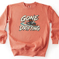 Gone Drifting Comfort Colors crewneck sweatshirt in Terracotta, laid flat. A rider in an orange jacket and helmet drifts a teal and white snowmobile, kicking up a teal snow trail, between the words Gone Drifting in cream brush lettering. Shown on a white background.
