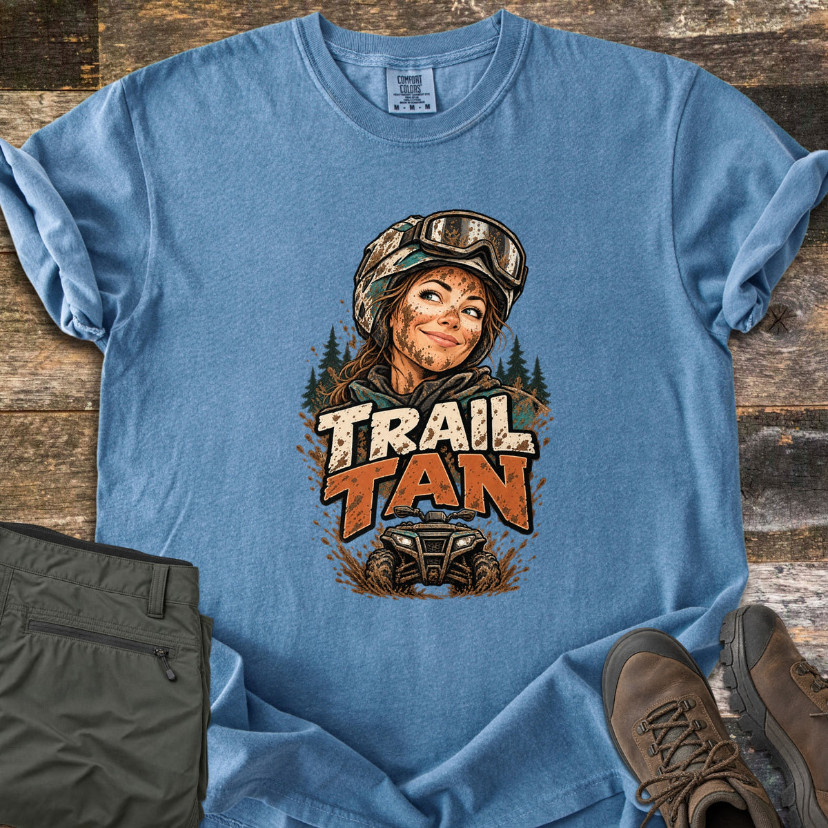 Trail Tan Comfort Colors T-shirt in Blue Jean, laid flat. Front print of a smiling woman in a mud-splattered helmet and goggles with pine trees, above the words Trail Tan and a muddy ATV. Olive pants and brown hiking boots sit beside it on weathered wood.