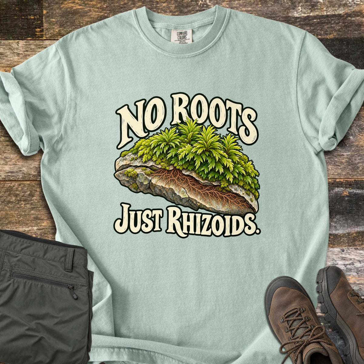 No Roots Just Rhizoids Comfort Colors T-shirt in Bay, laid flat. Green moss sprouts from a cracked rock with brown rhizoids threading underneath, between the words No Roots Just Rhizoids. On rustic wood beside folded olive hiking pants and brown hiking boots.