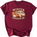 Winter Tucked In Comfort Colors T-shirt in Maroon, laid flat. A sleeping red fox curls into an oval that frames a snowy field with bare trees and a red barn, with WINTER arched above and TUCKED IN below in cream letters. The shirt lies on a white surface with sleeves cuffed.