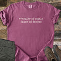 Fixer of Fences Comfort Colors T-shirt in Berry, laid flat. Off-white typewriter-style text across the chest reads wrangler of cattle, fixer of fences. The shirt has rolled sleeves and lies on weathered wood planks beside folded olive green pants and brown hiking boots.
