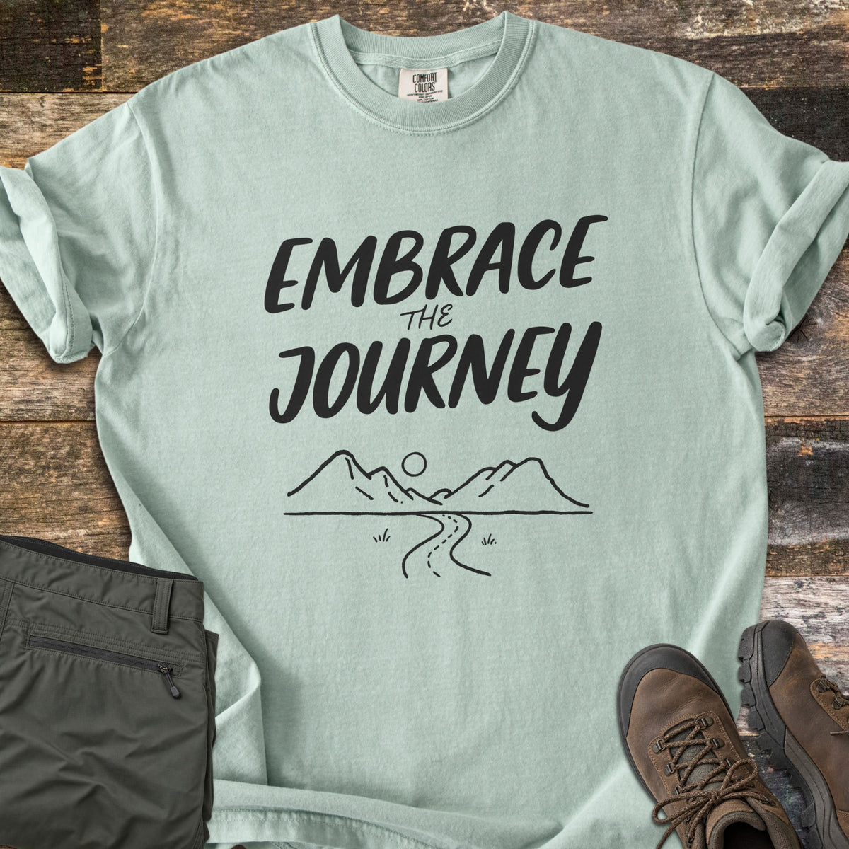 Embrace the Journey Comfort Colors T-shirt in Bay, laid flat. Black brush lettering reads Embrace the Journey above a line drawing of mountains, a sun, and a winding road. Sleeves cuffed on weathered wood beside folded green pants and brown hiking boots.