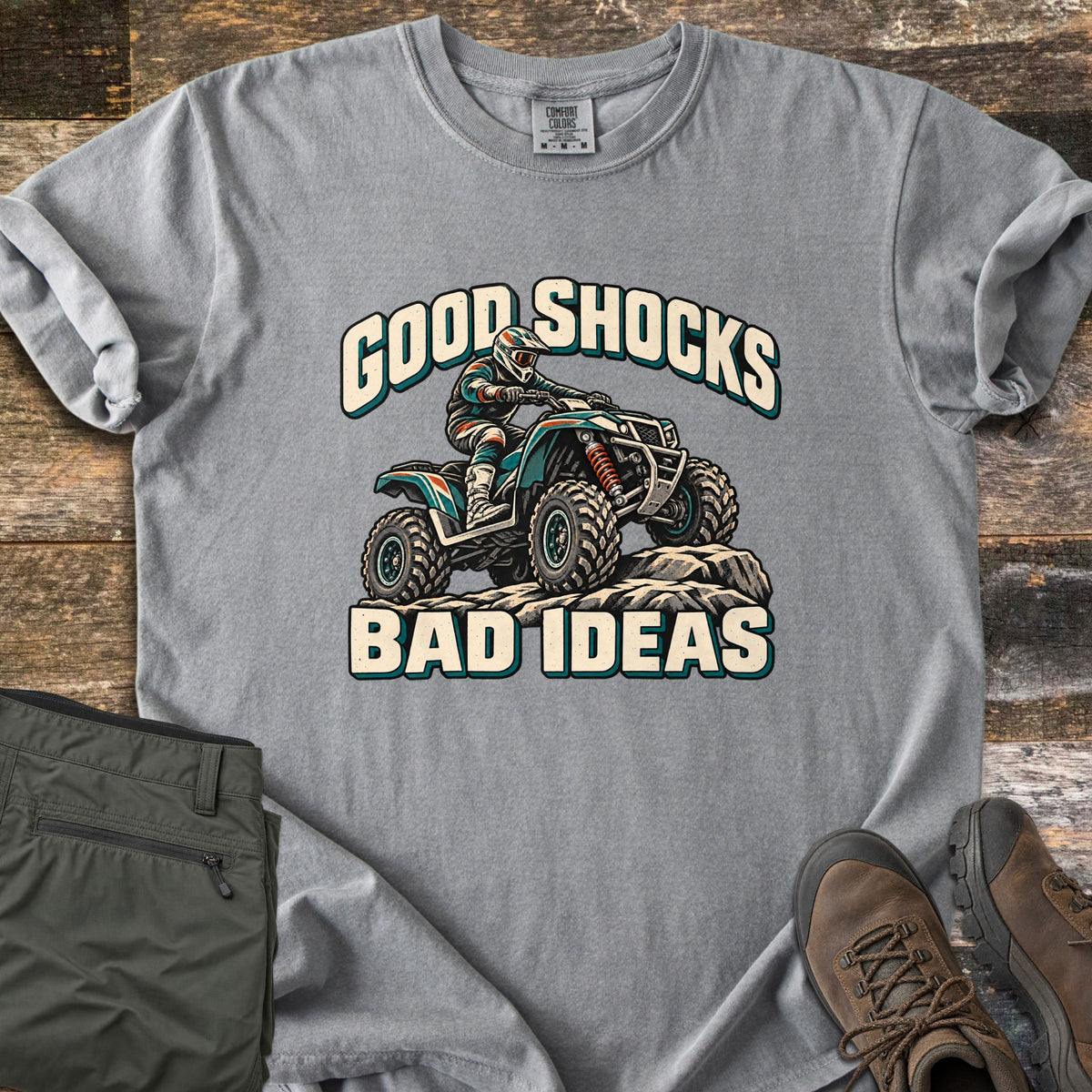 Good Shocks Bad Ideas Comfort Colors T-shirt in Grey, laid flat. An illustrated rider in a helmet and goggles climbs rocks on a teal ATV between the words Good Shocks and Bad Ideas. It lies on weathered wood beside folded green cargo pants and brown hiking boots.